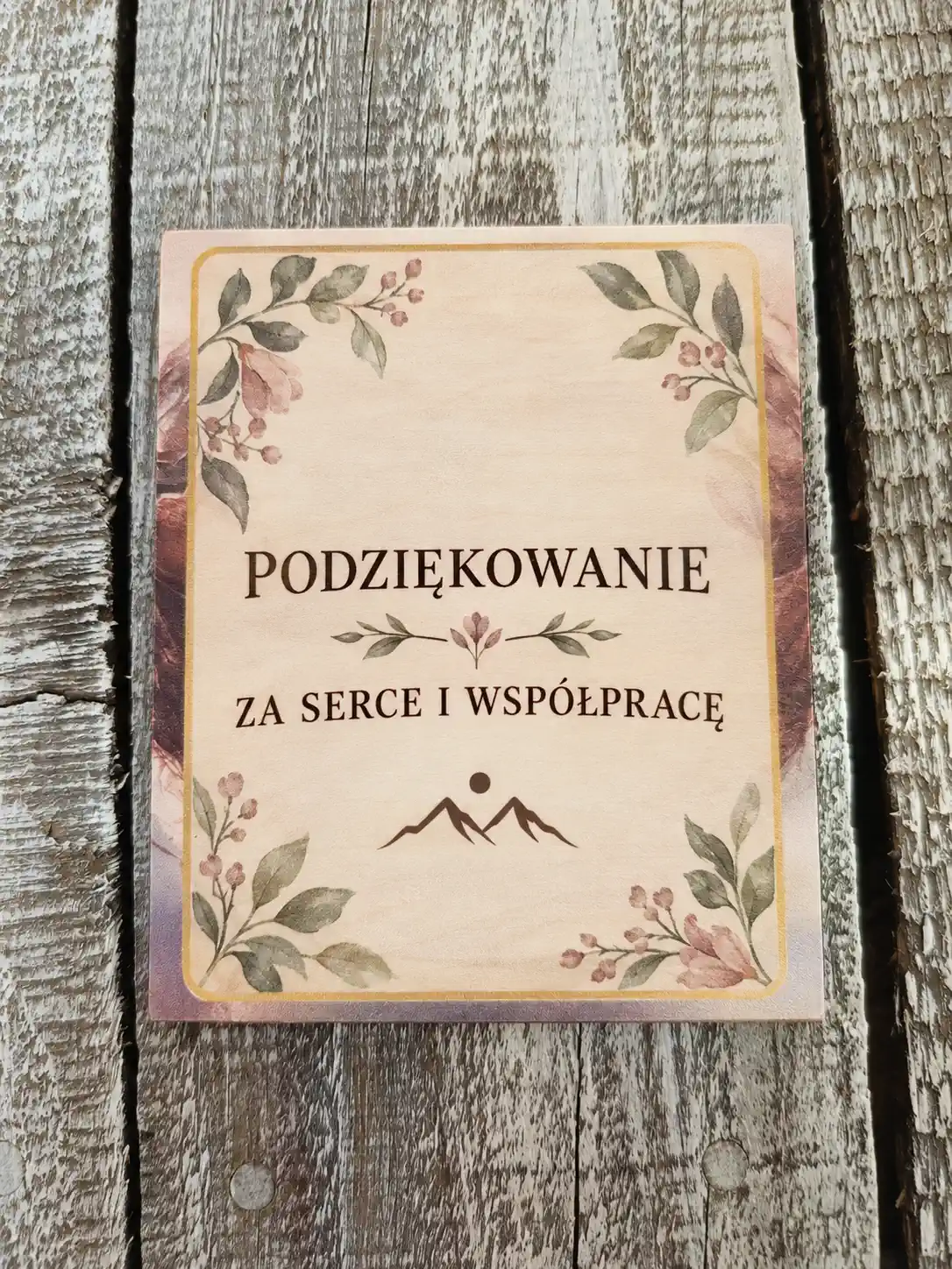 Drewniana tabliczka z pełnokolorowym nadrukiem kwiatowym i ciemnym napisem, leżąca na szarych, zwietrzałych deskach