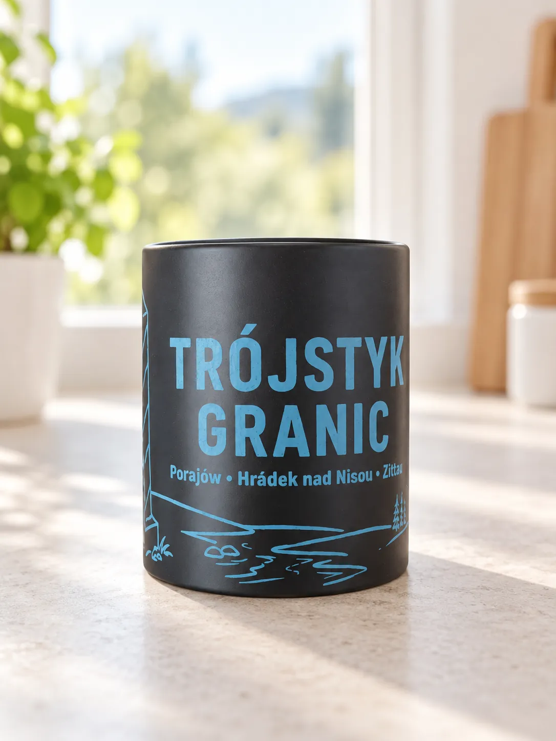 Czarny matowy kubek ceramiczny z niebieskim nadrukiem: napis i rysunek liniowy jeziora, na jasnym blacie.