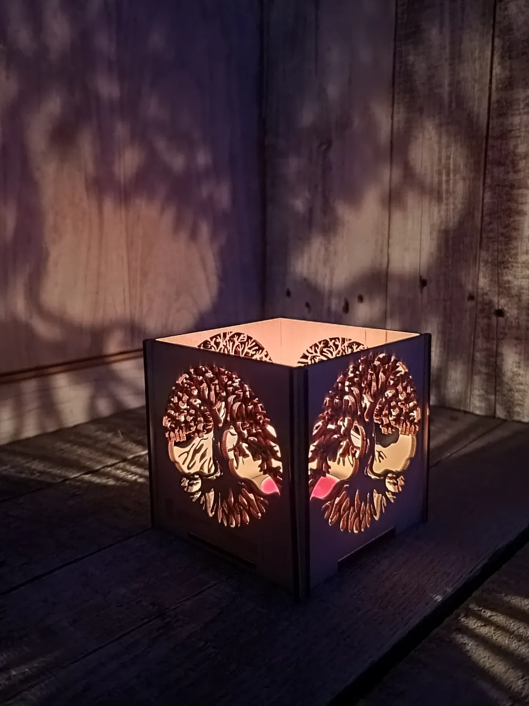 Lampion sześcienny ze sklejki z ażurową rozetą drzewa życia, zapalony na drewnianych deskach, z cieniem wzoru na ścianie.